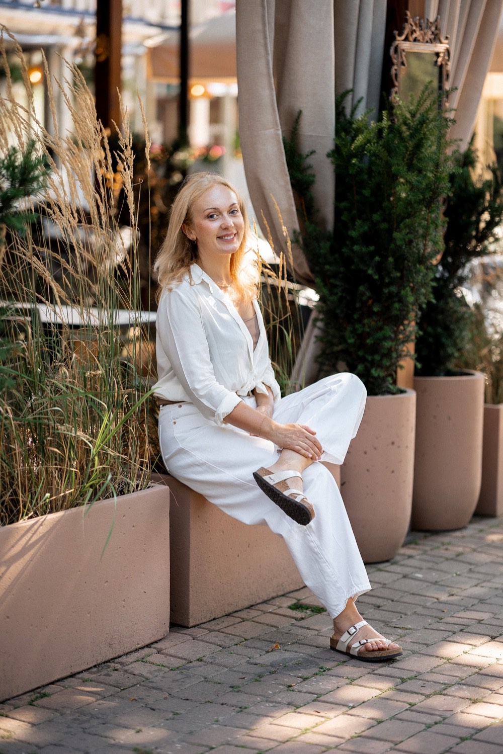 Svitlana sitting outdoors on a stone planter in a white outfit, relaxed and smiling